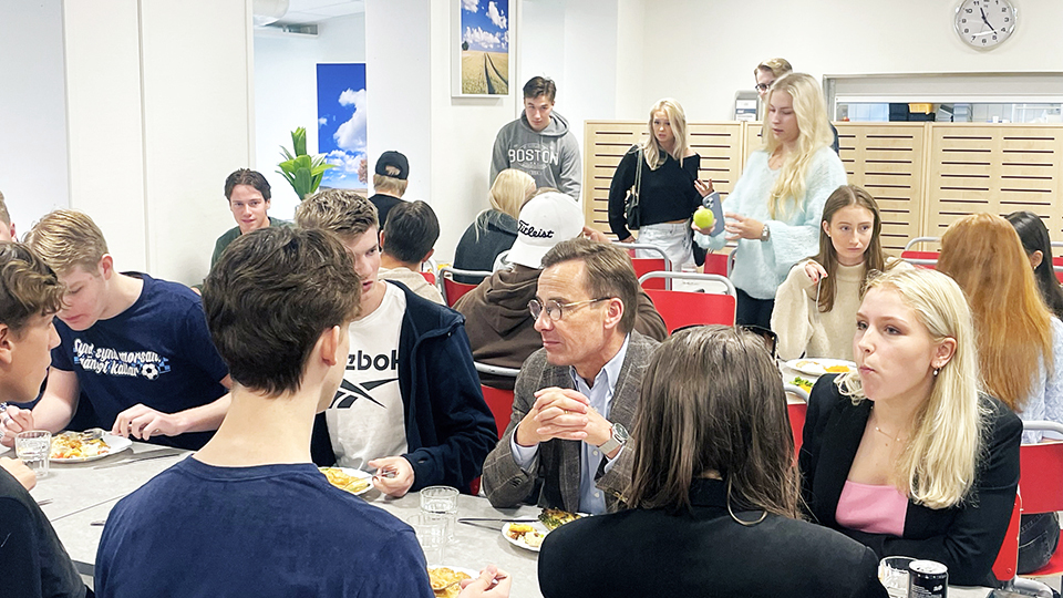 Bilden visar statsministern som sitter vid ett bord i matsalen med elever från Österåkers gymnasium och äter lunch